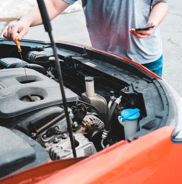 Man inspecting car engine while holding phone outdoors, checking oil levels.
